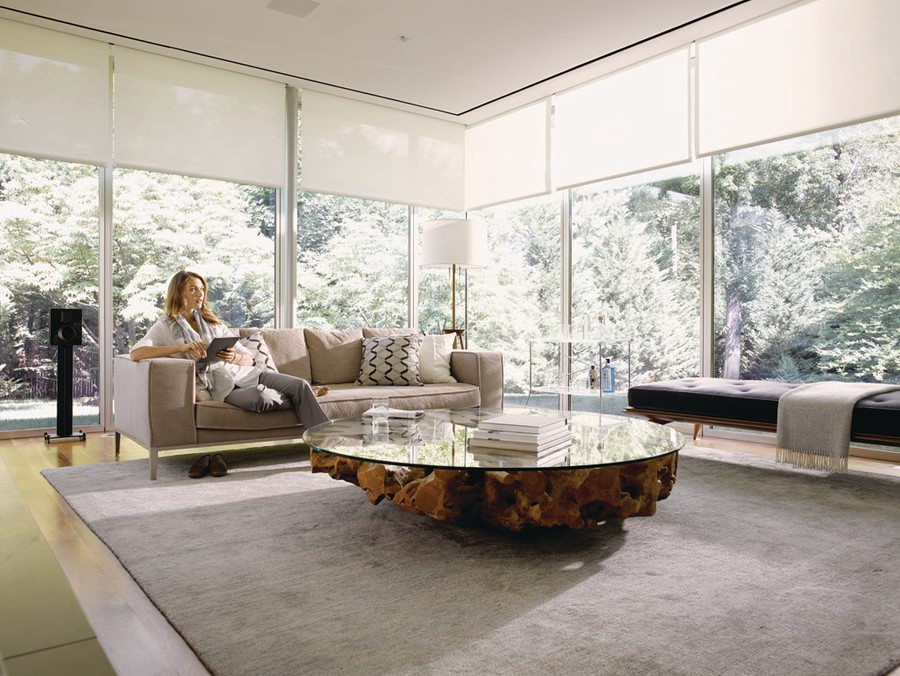 woman sits on living room couch while holding a tablet. Behind her are floor-to-ceiling windows with shades.