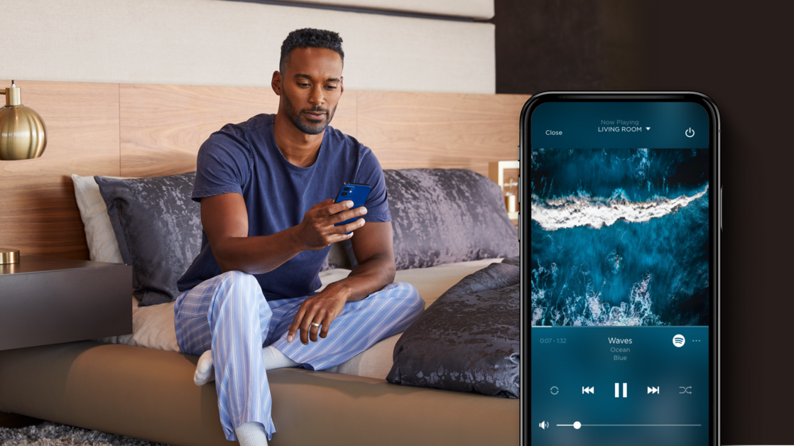 Man sitting on a bed using the Savant App with a close-up of the music interface displayed on the screen. 