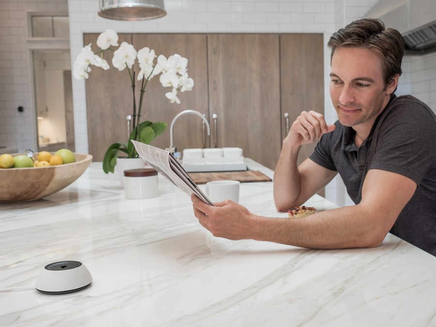 A man sitting at a marble kitchen island, reading a newspaper with a Josh micro placed on the countertop. The modern kitchen features a bowl of fresh fruit, a white orchid centerpiece, and wooden cabinetry in the background.