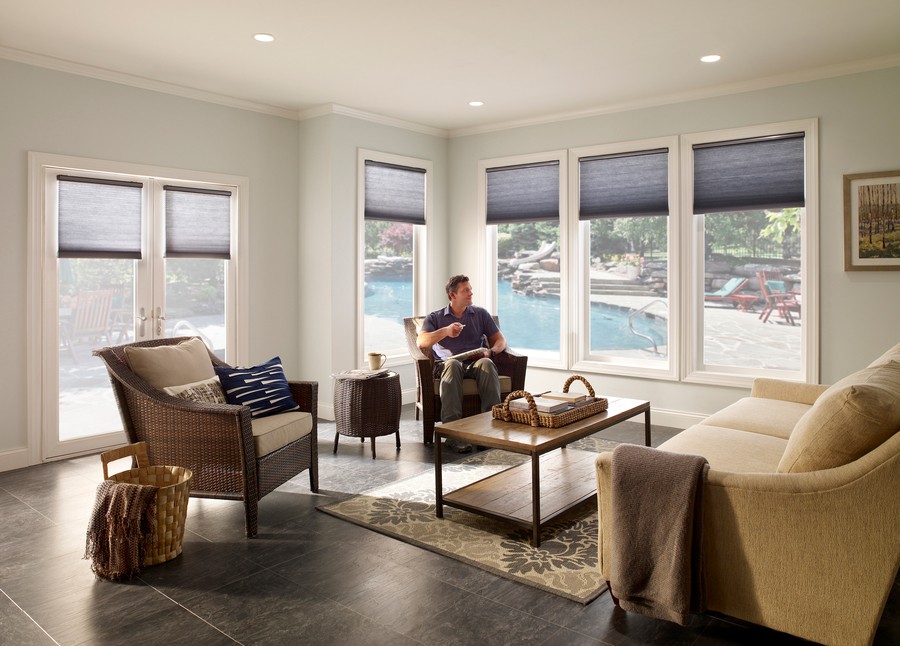 A man relaxes in a chair in a living area, outside the windows is a patio and swimming pool.